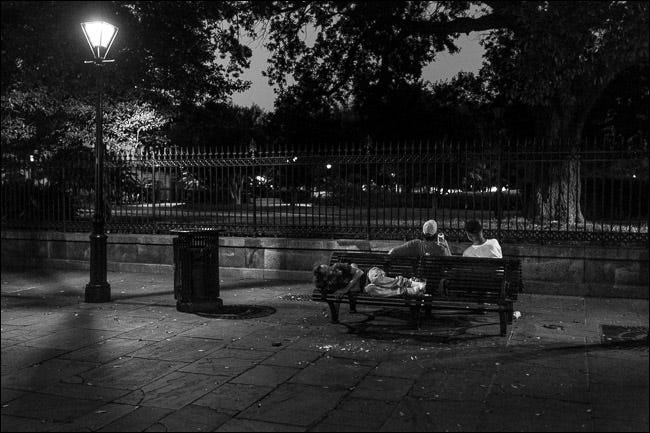zwei Männer sitzen nachts auf einer Parkbank;  Ein Mann schläft auf einem Sitz hinter ihnen